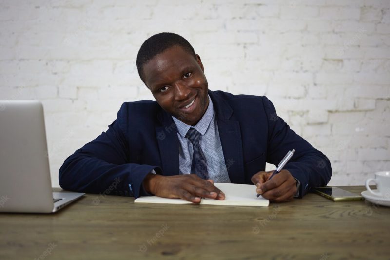 Picture of attractive African lawyer wearing suit working at his office desk, making notes in diary, looking and smiling . happy businessman writing down his ideas and plans in copybook Free Photo