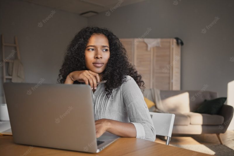 Picture of fashionable attractive young dark skinned woman freelancer in shirt sitting at workplace at home and using high speed wirless internet connection on laptop, having thoughtful look Free Photo