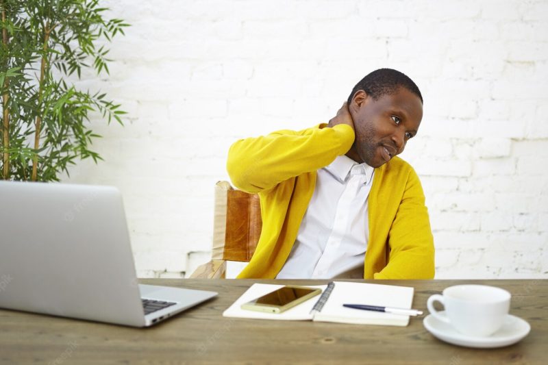 Picture of modern fashionable young dark skinned businessman rubbing neck, feeling frustrated and uncertain about something, sitting at workplace with open laptop, diary, mug and mobile phone on desk Free Photo