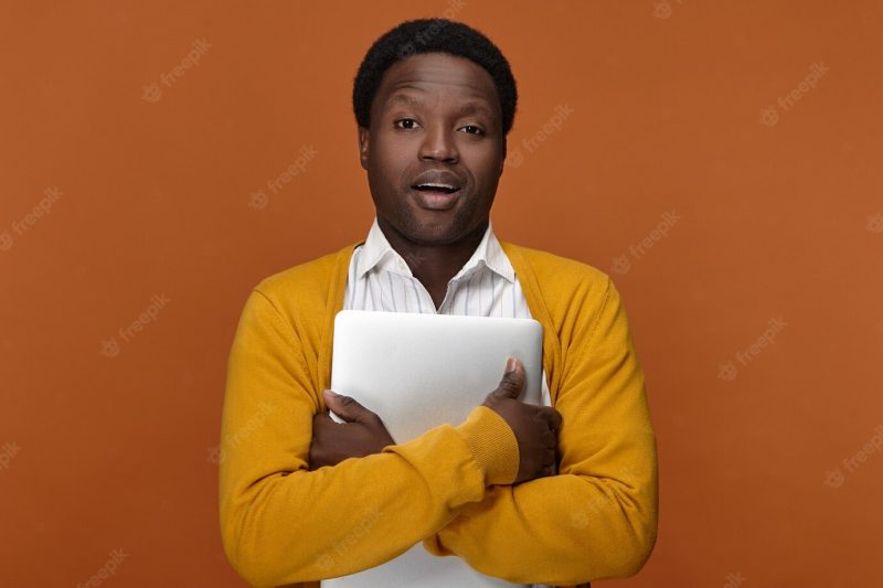 Picture of stylish young dark skinned manager in yellow cardigan holding generic laptop, leaving office after work. people, modern technology, job, occupation and electronic gadgets concept Free Photo