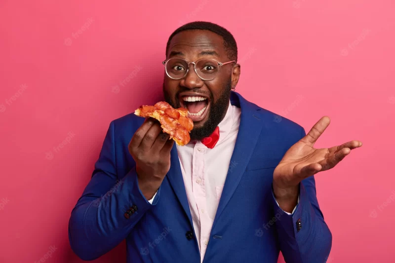 Pizza time concept. positive male office worker or entrpreneur in suit holds big piece of pizza, raises palm, has good appetite Free Photo