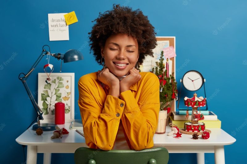 Pleasant looking woman sits with closed eyes, toothy smile on chair, has good dreams, enjoys domestic atmosphere, poses against white desk in study room dressed in yellow jacket keeps hands under chin Free Photo