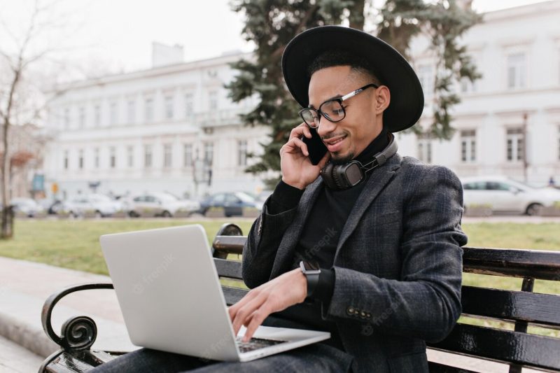 Pleased african freelancer talking on phone and typing on keyboard. outdoor photo of international student in black outfit using laptop on nature. Free Photo