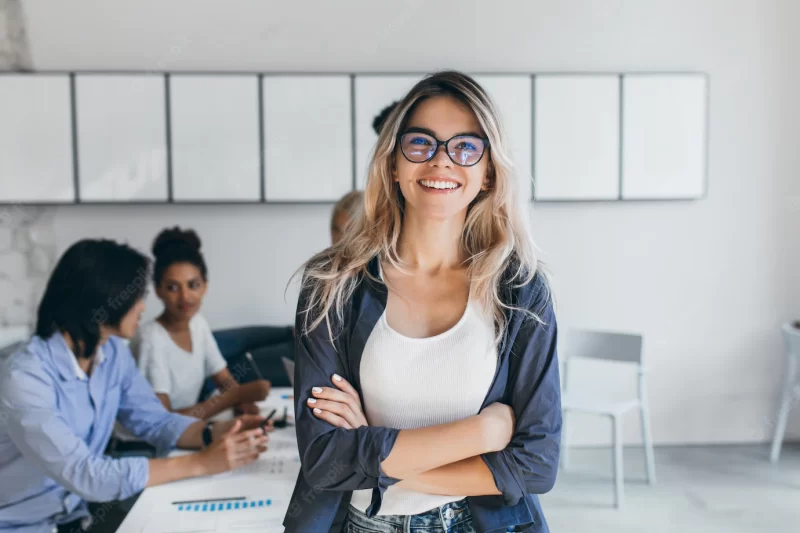 Pleased female secretary in trendy glasses posing in office after meeting with colleagues. indoor portrait of stylish businesswoman with Asian and African workers. Free Photo