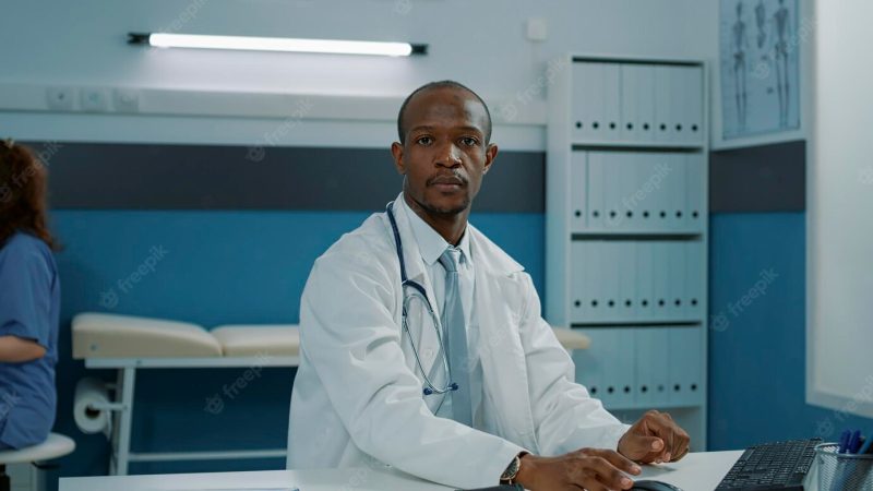 Portrait of african american doctor using computer in cabinet, working on health support. general practitioner wearing white coat and stethoscope, feeling confident about service and expertise. Free Photo