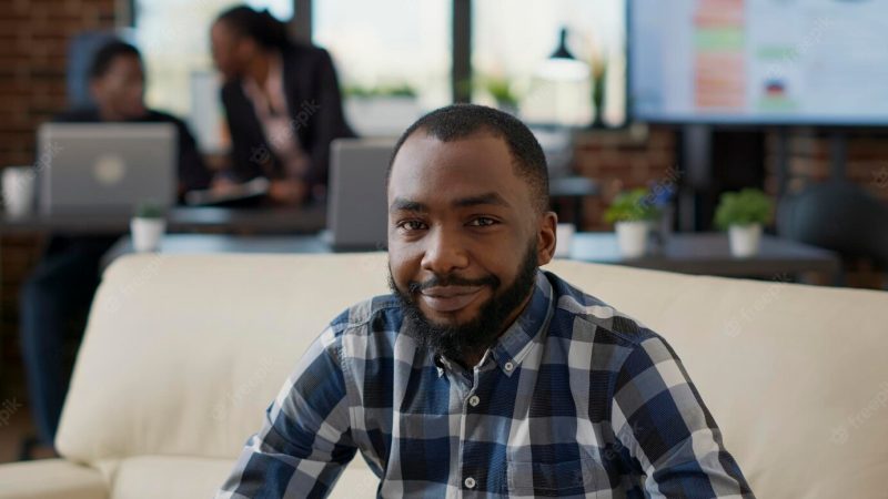 Portrait of african american person smiling in office, sitting on couch to work on laptop. company employee using financial statistics to create job development and business growth. Free Photo