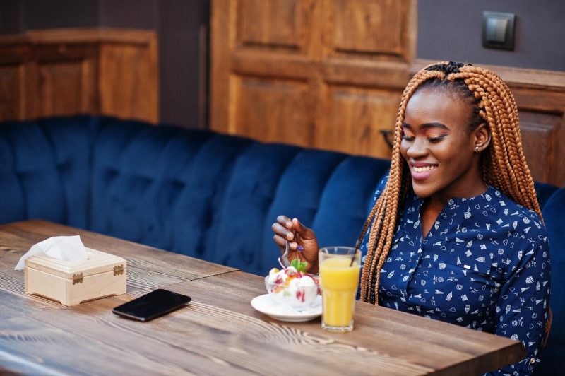 Portrait of beautiful young african business woman with dreadlocks wearing blue blouse and skirt sitting in cafe with ice cream and pineapple juice Free Photo