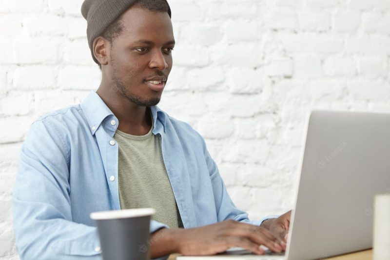 Portrait of black male in stylish clothes using free internet connection at cafeteria, working at laptop, surfing social networks and drinking coffee. businessman working with modern device at cafe Free Photo