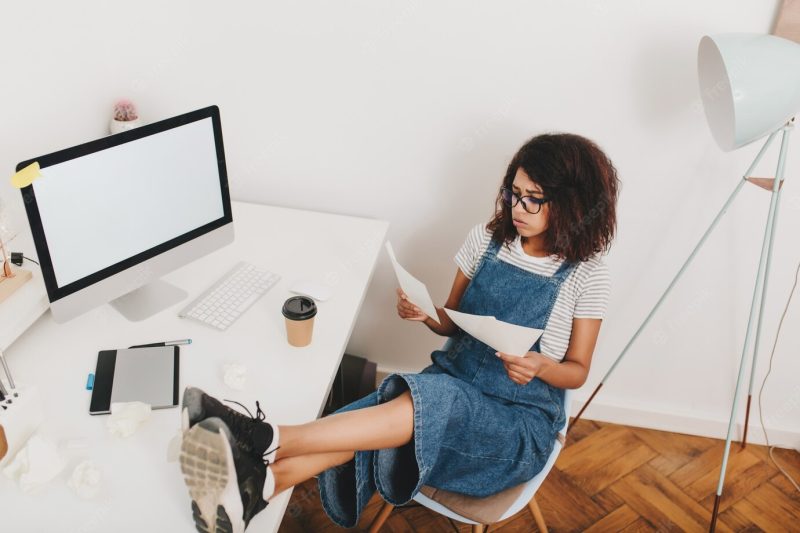 Portrait from above of busy young woman in glasses sitting with legs on table beside computer and tablet Free Photo