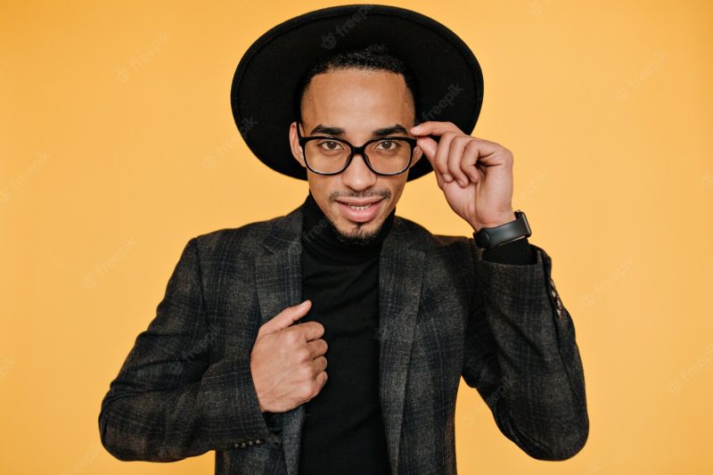 Portrait of handsome brown-eyed male model playfully touching his hat. indoor photo of well-dressed African man in good mood. Free Photo