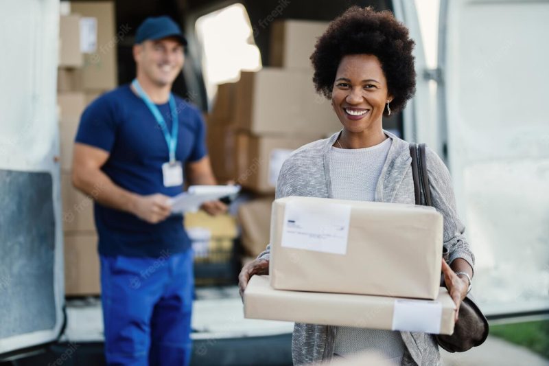 Portrait of happy black woman feeling satisfied with package delivery ad holding boxes while looking at camera the courier is in the background Free Photo