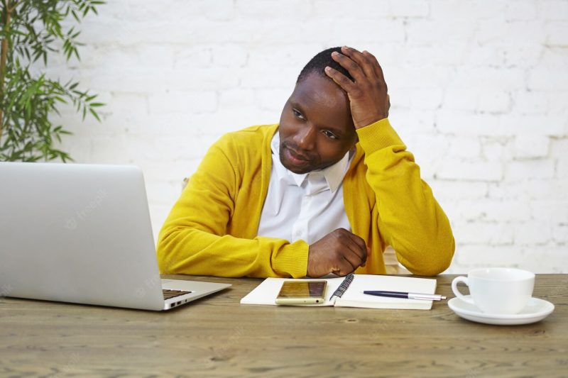 Portrait of sad afro american male employee wearing yellow cardigan touching head feeling tired and overworked because of stress or failure at work, sitting at desk with laptop, coffee and diary Free Photo