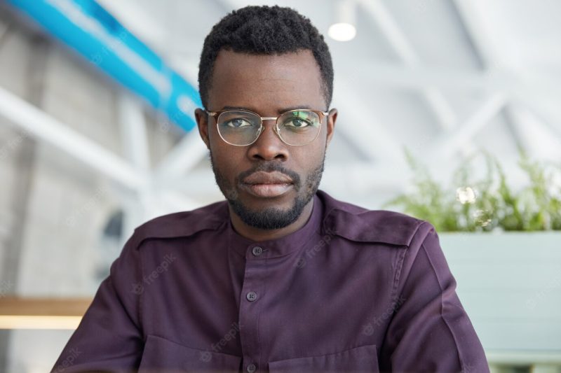 Portrait of serious confident male office worker in spectacles and formal shirt, with dark skin, poses in spacious cabinet Free Photo