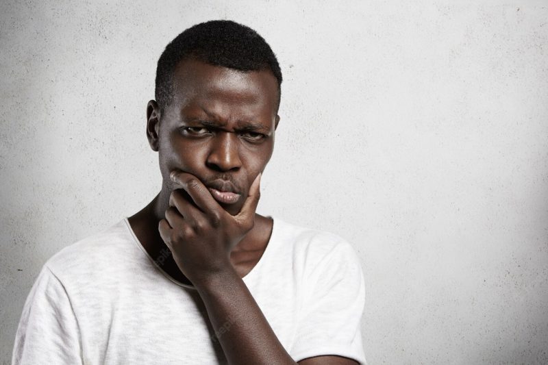 Portrait of skeptical African young man looking with suspicious or annoyed expression, holding hand on chin, doubting, thinking over something. Free Photo