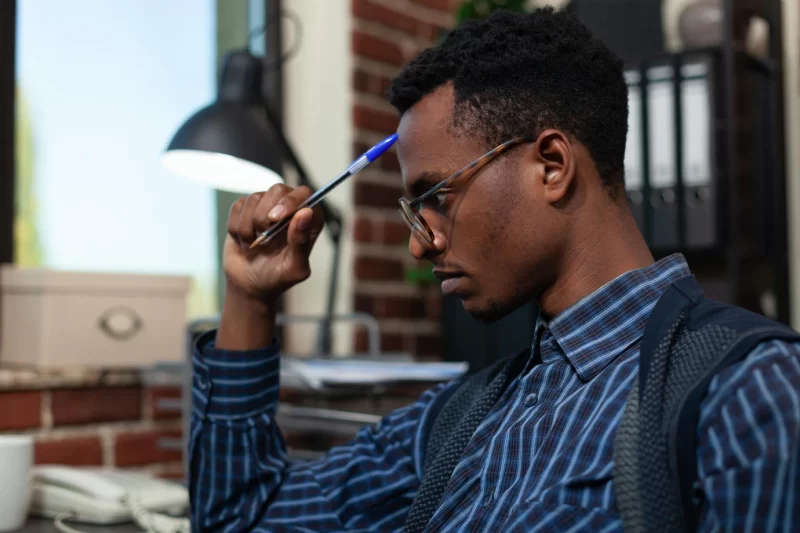 Portrait of startup employee touching forehead with pen looking focused at laptop screen with business analyitics. african american with glasses thinking about sales results sitting at desk. Free Photo