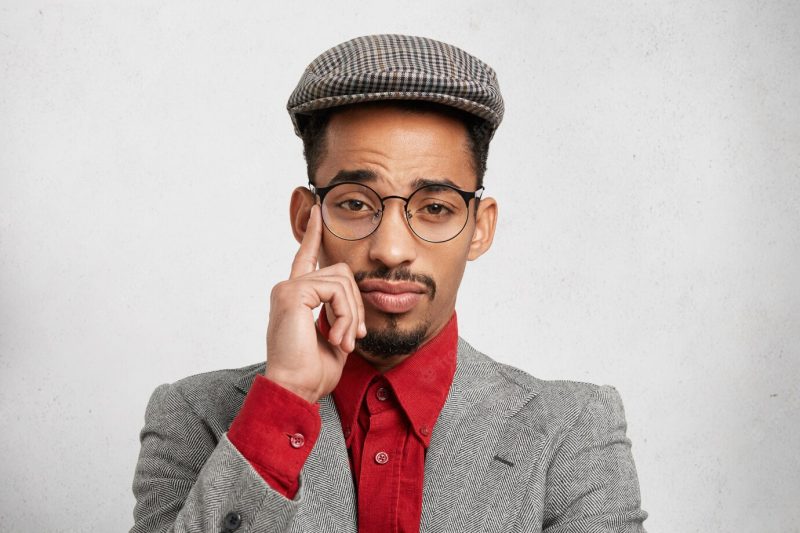 Portrait of thoughtful pensive mixed race male employee wears trendy cap, red shirt and jacket, has wise expression Free Photo