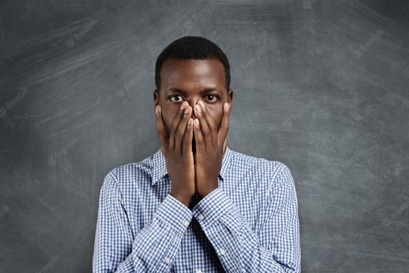 Portrait of young Afro-American man in checkered shirt covering his mouth with both hands and looking with shocked and guilty expression as if he did something wrong, standing at blank chalkboard Free Photo