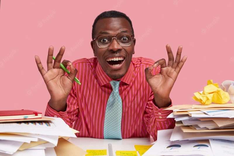 Positive Afro American man makes okay gesture with both hands, smiles broadly, demonstrates everything is alright, dressed in formal clothes, holds pen Free Photo