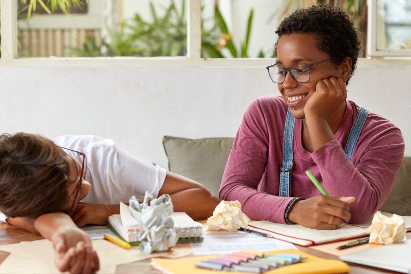 Positive black boyish woman looks at groupmate who feels tired, prepare for upcoming exam together, make notes of creative ideas, surrounded with crag of paper. team working and studying concept Free Photo