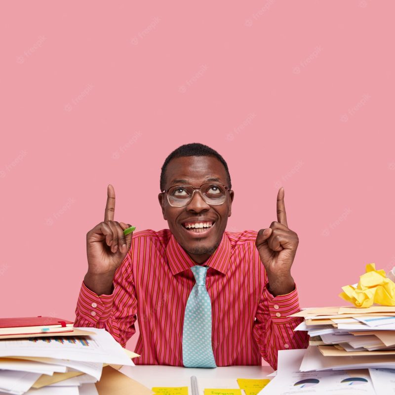 Positive black business owner points with both index fingers upwards, satisfied with successfully signed agreement, dressed formally, sits at desktop Free Photo