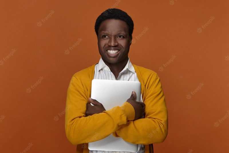 Positive talented young male freelancer of african appearance carrying portable computer, having happy cheerful facial expression, smiling broadly, enjoying his high speed electronic device Free Photo