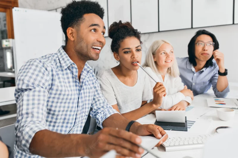 Pretty african female student biting pencil while thinking about something. indoor portrait of pleased black office worker in checkered blue shirt sitting at the table with colleagues. Free Photo