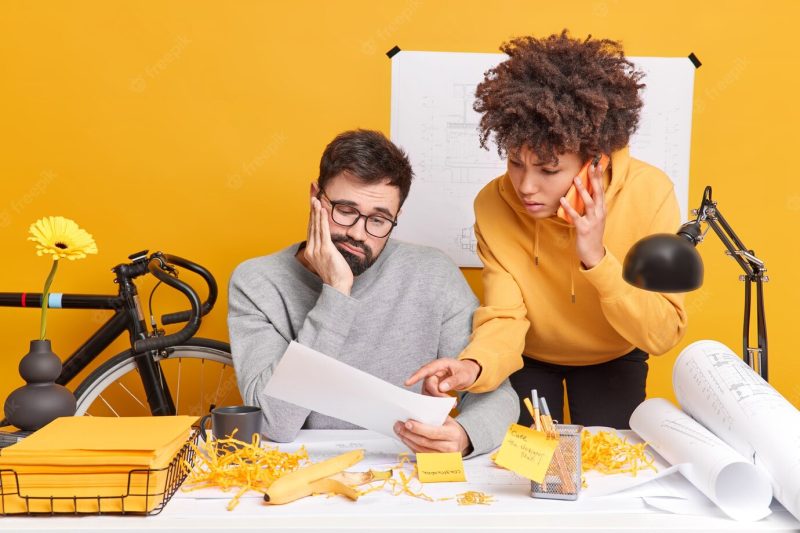 Professional male engineer looks sadly at paper listens explanation from female trainee who talks via smartphone pose at coworking space together. construction workers work on technical plans Free Photo Professional male engineer looks sadly at paper listens explanation from female trainee who talks via smartphone pose at coworking space together. construction workers work on technical plans Free Photo