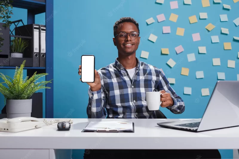 Project manager pointing phone device with mockup display to camera. young adult business person sitting in company modern workspace office while showing white screen isolated template to camera. Free Photo