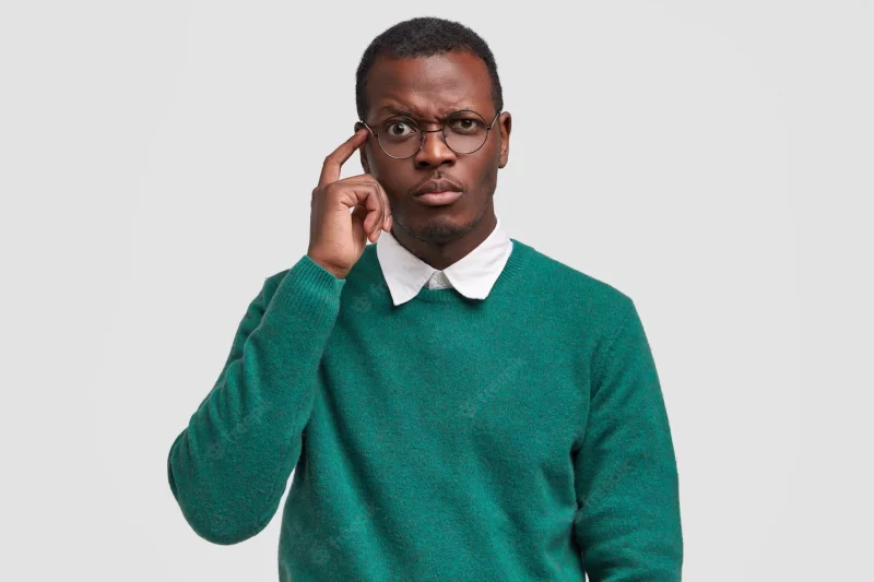 Puzzled dark skinned man frowns face, keeps index finger on temple, tries to concentrate, being deep in thoughts, wears green jumper Free Photo