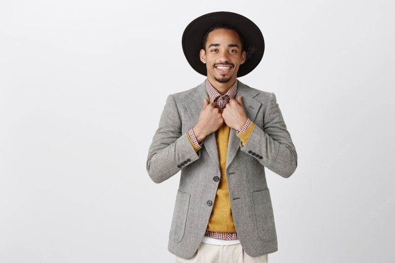 Ready to impress all girls in club. indoor shot of attractive confident dark-skinned male model in stylish outfit and black hat, touching jacket while choosing new clothes in shop over gray wall Free Photo