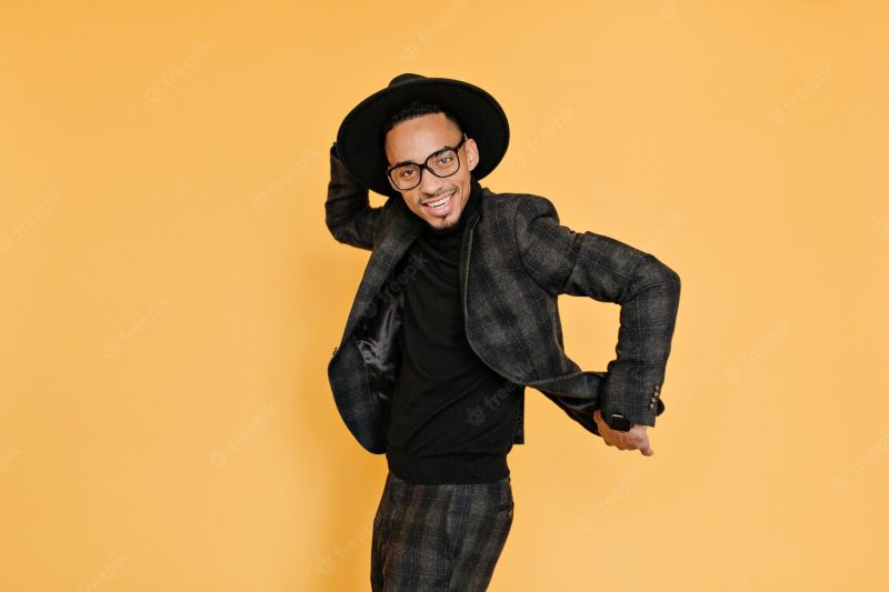 Refined african man having fun during photoshoot . indoor photo of smiling tall black guy in hat and glasses dancing on yellow wall. Free Photo