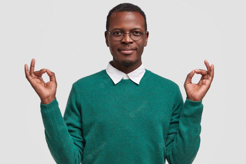 Relaxed dark skinned male worker rests after making task, keeps both hands in okay gesture, meditates indoor Free Photo