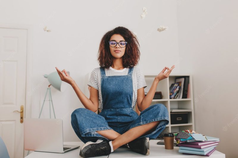 Relaxed young woman with bronze skin sitting in lotus pose on the table with laptop and documents Free Photo