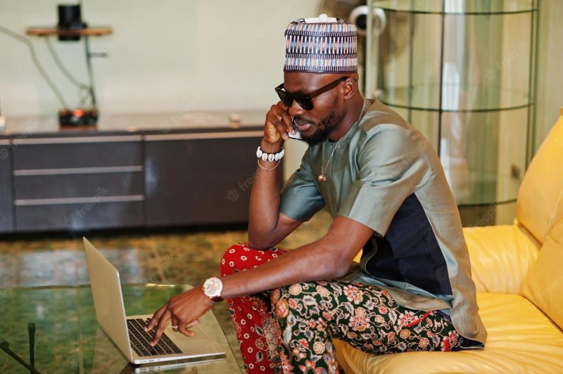 Rich african man sitting on sofa at his appartment and working on notebook and speaking on phone Free Photo