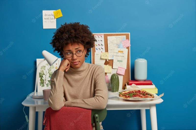 Sad woman has afro haircut sits at chair, wears round spectacles and beige jumper, sits in coworking space, table with notes desk lamp and delicious pizza behind. Free Photo