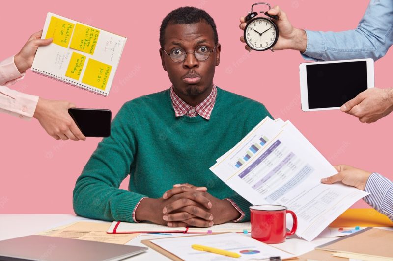 Sad young African American man dressed in green sweater, sits at desktop, hands with papers, alarm clock, touch pad, notepad with stickers Free Photo