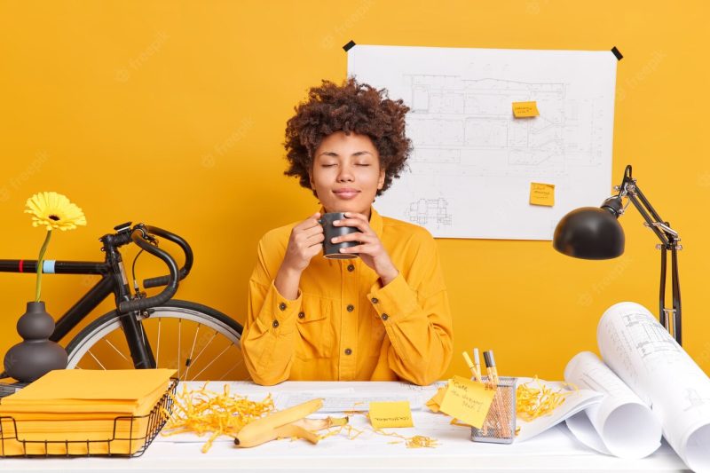 Satisfied relaxed woman architect office worker has coffee break closes eyes from pleasure as smells aromantic beverage draws draft at desktop creates sketches Free Photo