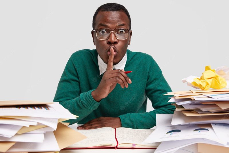 Secret African American male CEO shows silence sign, works on task received from boss, writes down ideas in notebook, has surprised facial expression Free Photo