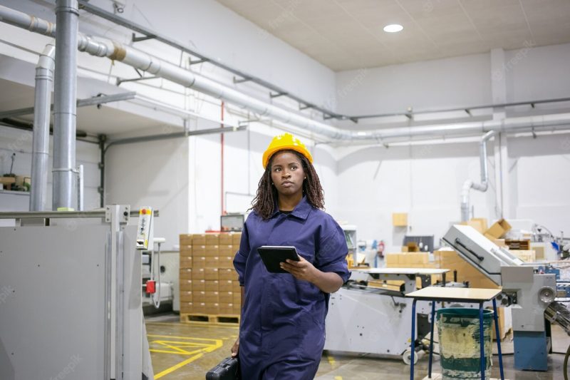 Serious african american female worker in protective uniform walking to workplace on plant floor, holding tablet and case with tools Free Photo