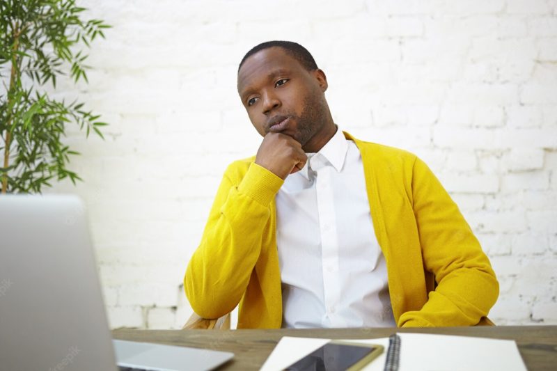 Serious african american male employee dressed stylishly sitting at white brick wall at his workplace, using generic portable computer, rubbing his chin, having thoughtful facial expression Free Photo