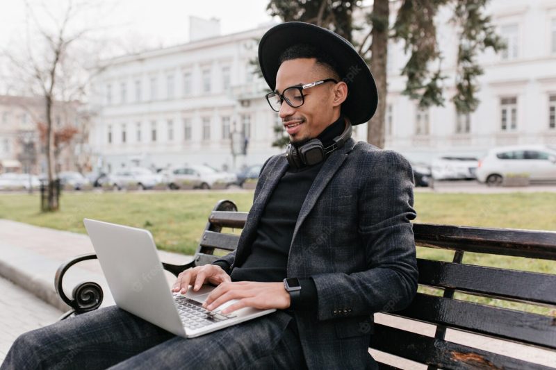 Serious african man in black shirt and pants using laptop under open sky. outdoor photo of mulatto freelancer resting on park bench. Free Photo