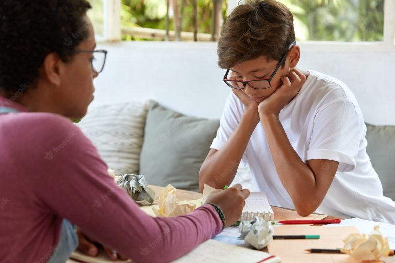 Serious concentrated asian boy keeps hands on cheeks, wears transparent glasses, focused in notepad, listens explanation from tutor, sit opposite each other in terrace cafe, solve problematic task Free Photo