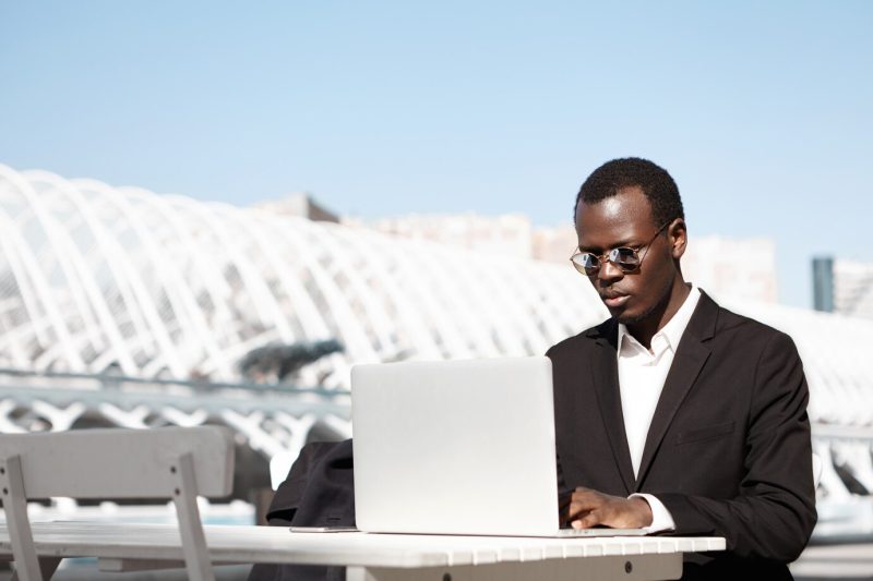 Serious confident black entrepreneur in round shades and formal suit looking at screen of laptop in front of him with concentrated expression, waiting for business partners for meeting at urban cafe Free Photo