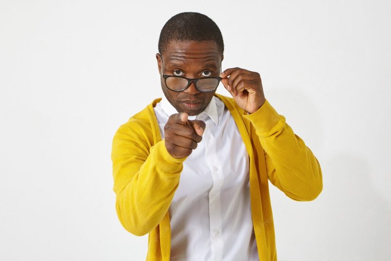 Serious confident dark skinned male teacher in eyewear pointing index finger , having strict look, warning his students, standing isolated at white wall with copy space for your text Free Photo