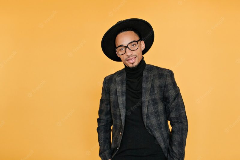 Serious dark-eyed mulatto guy standing on yellow wall with hands in pockets. indoor photo of african young man in casual suit wears black hat. Free Photo