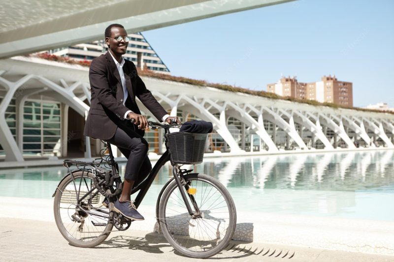 Serious dark-skinned european entrepreneur wearing elegant black suit and mirror sunglasses standing outdoors with his bicycle while waiting for partner for lunch, messaging him on smartphone Free Photo