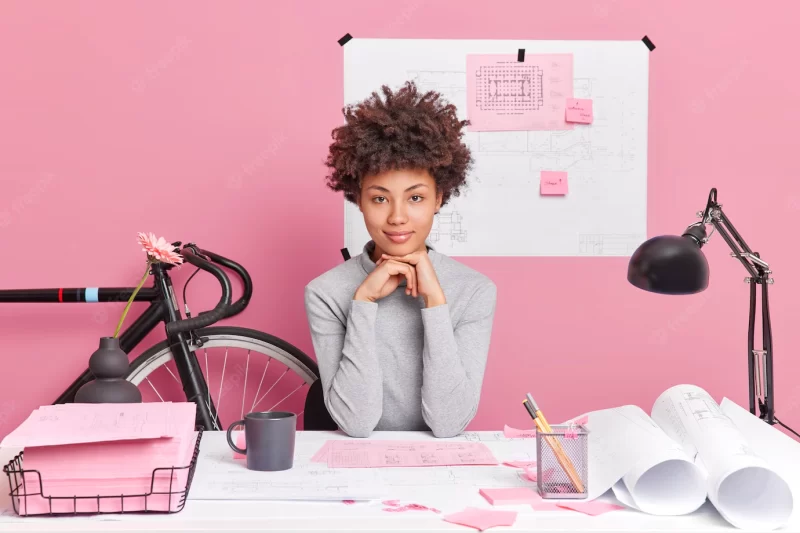 Serious dark skinned female office worker prepares construction project or interior designer poses at desktop with papers blueprints in office Free Photo