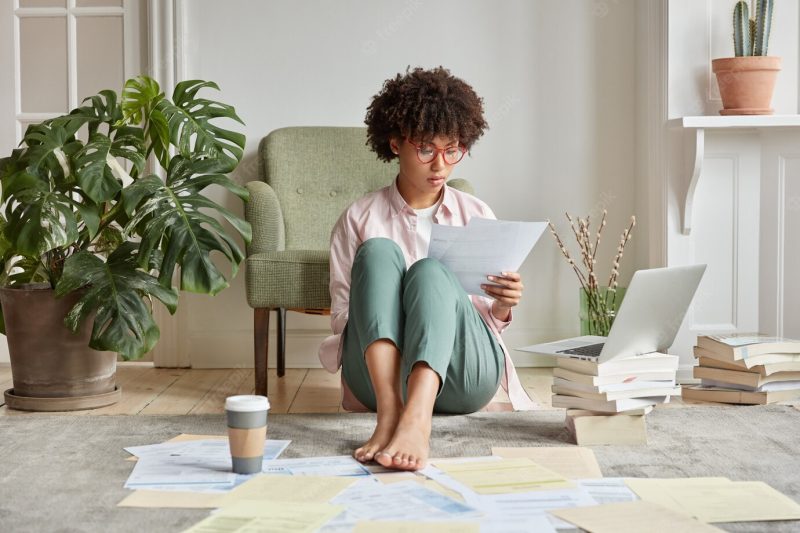 Serious dark skinned female worker hipster waits for update installation on laptop computer, sits on floor with many papers Free Photo