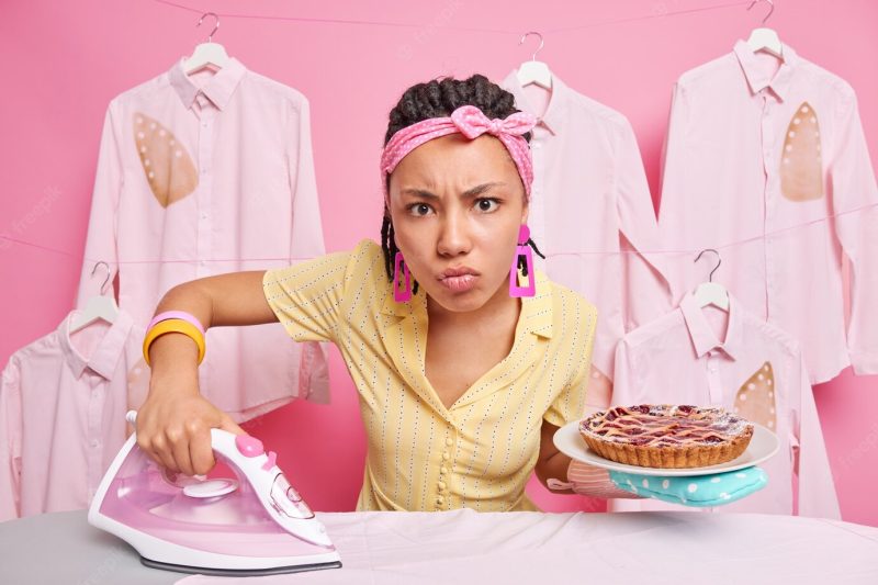 Serious housewife busy cooking and ironing at home holds delicious baked pie and electric iron poses near ironing board dressed in domestic clothes busy doing household chores has annoyed expression Free Photo