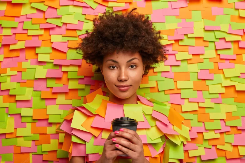 Serious looking ethnic office worker has coffee break, holds takeout beverage in hands, looks directly at camera, talks with colleague, poses indoor against adhesive notes on wall. people, drinking Free Photo